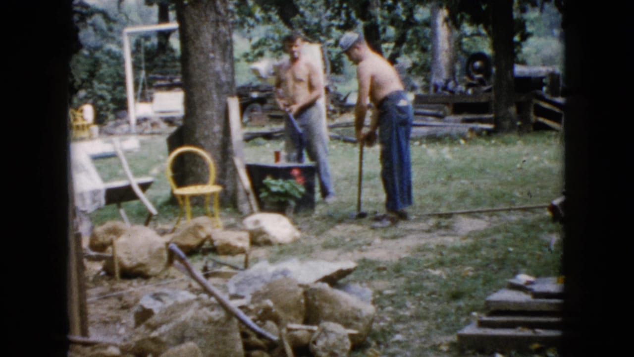 In Wauconda, Illinois home-movie footage from 1960, two shirtless men stand in a yard beside rocks, trees, a yellow chair, and work materials.