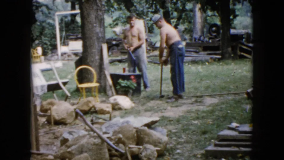In Wauconda, Illinois home-movie footage from 1960, two shirtless men stand in a yard beside rocks, trees, a yellow chair, and work materials.