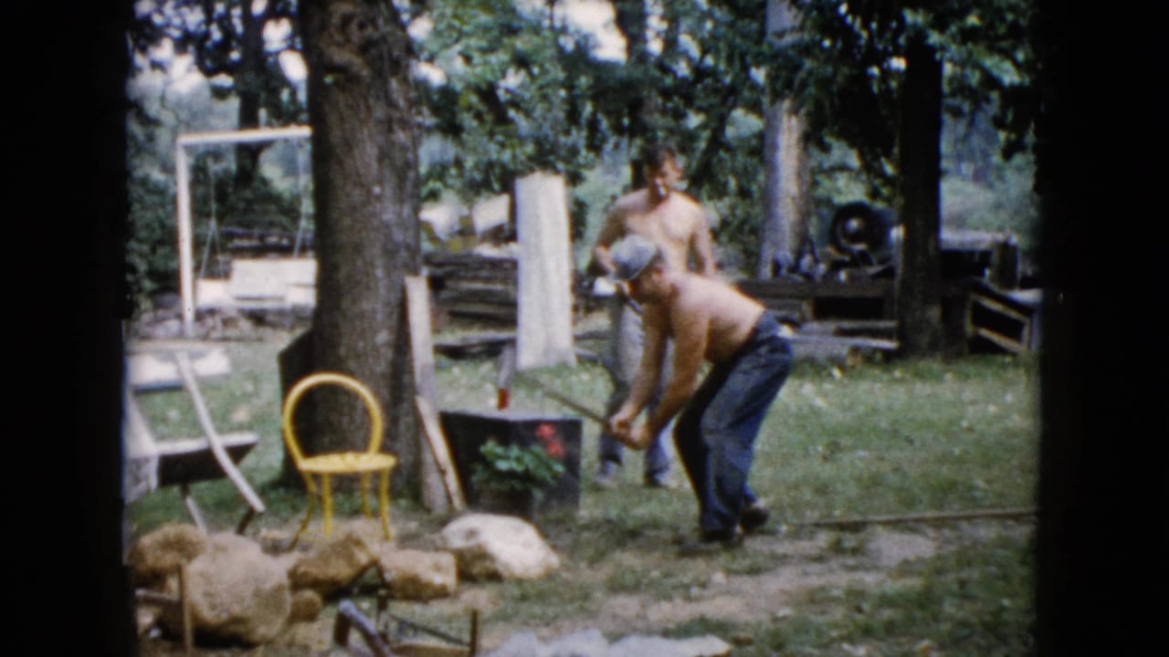 A shirtless man bends with a tool near rocks while another man stands behind him in the yard.