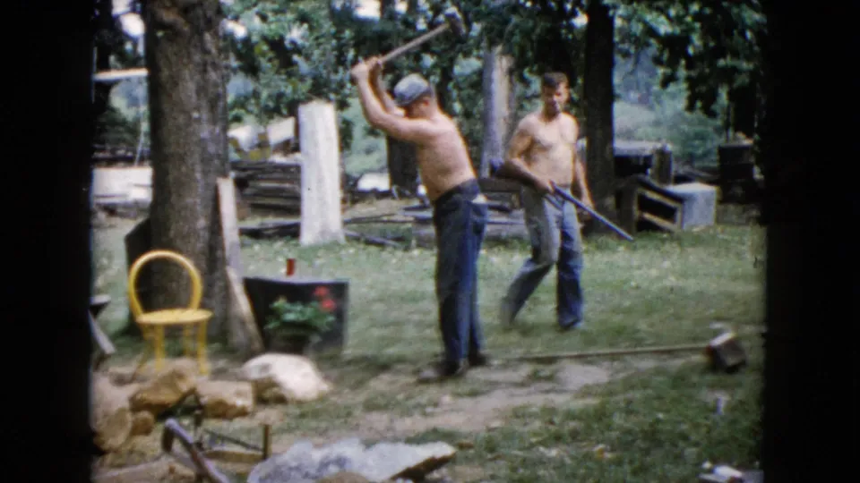 In the 1960 Wauconda yard footage, one man raises a hammer above the rocks while the other holds a long tool nearby.