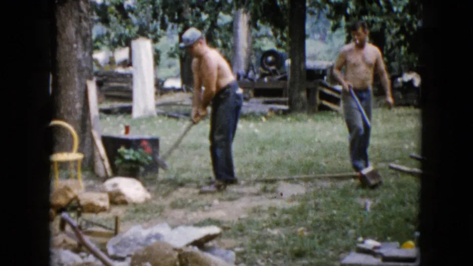 The clip ends with two shirtless men in a 1960 Wauconda backyard, surrounded by tools, stones, trees, grass, and a yellow chair.