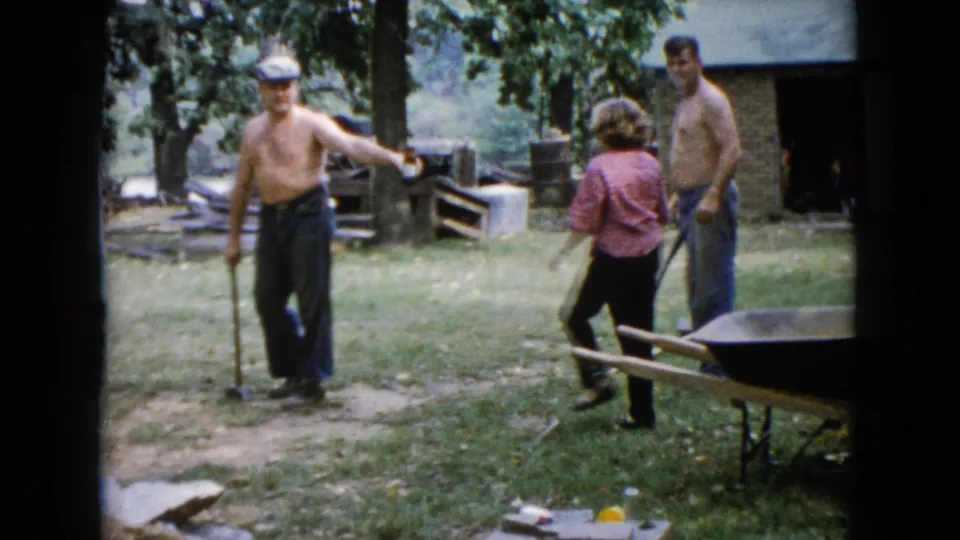 The backyard work scene continues as the man with the sledgehammer stands near a path while two adults reposition nearby.
