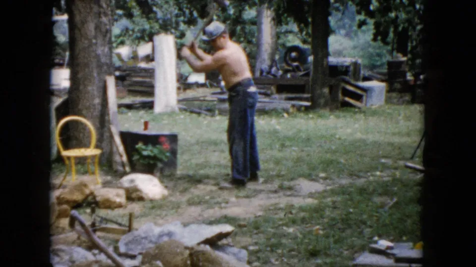 The man raises the sledgehammer for another swing in the 1960 Wauconda backyard, with a yellow chair, rocks, and yard materials nearby.