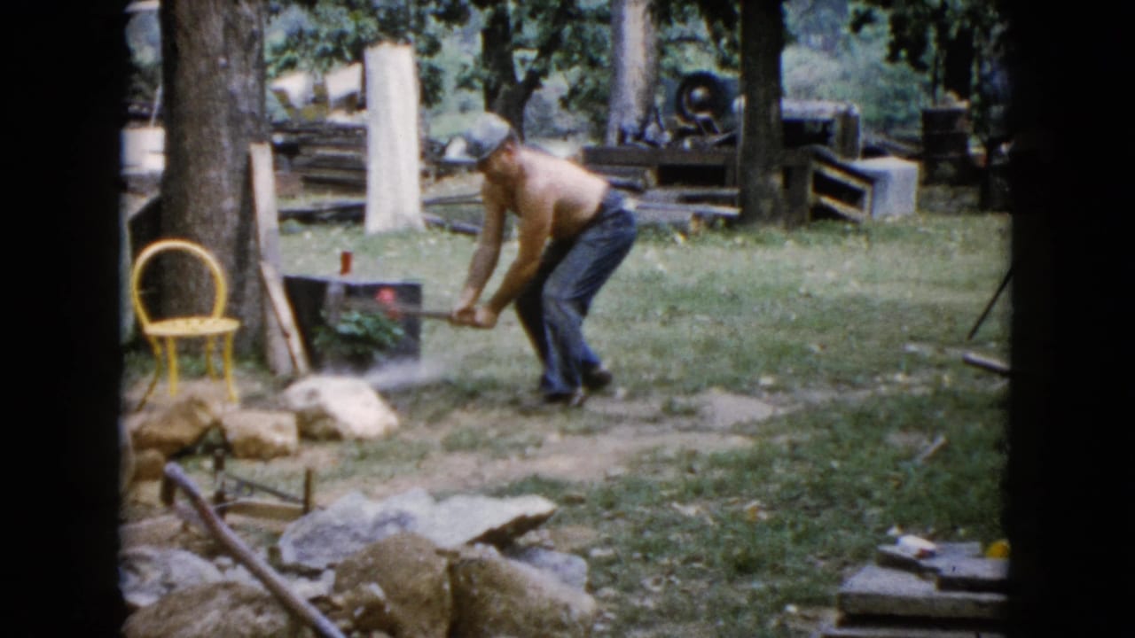 Shirtless man bends toward a rock with a hammer in a grassy yard.