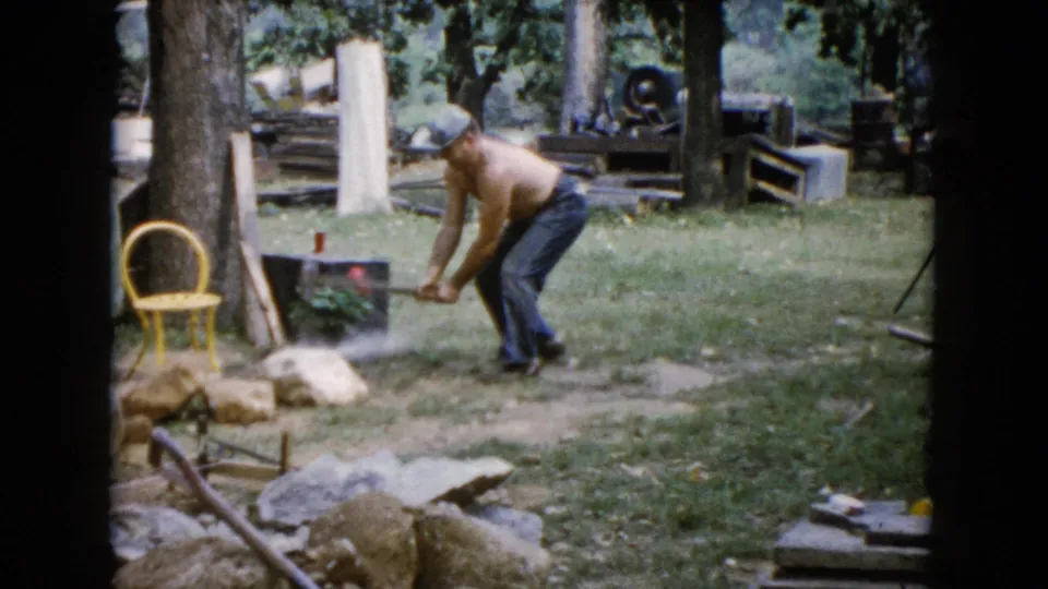 In 1960 Wauconda, Illinois home-movie footage, a shirtless man works over a rock with a hammer in a grassy yard.