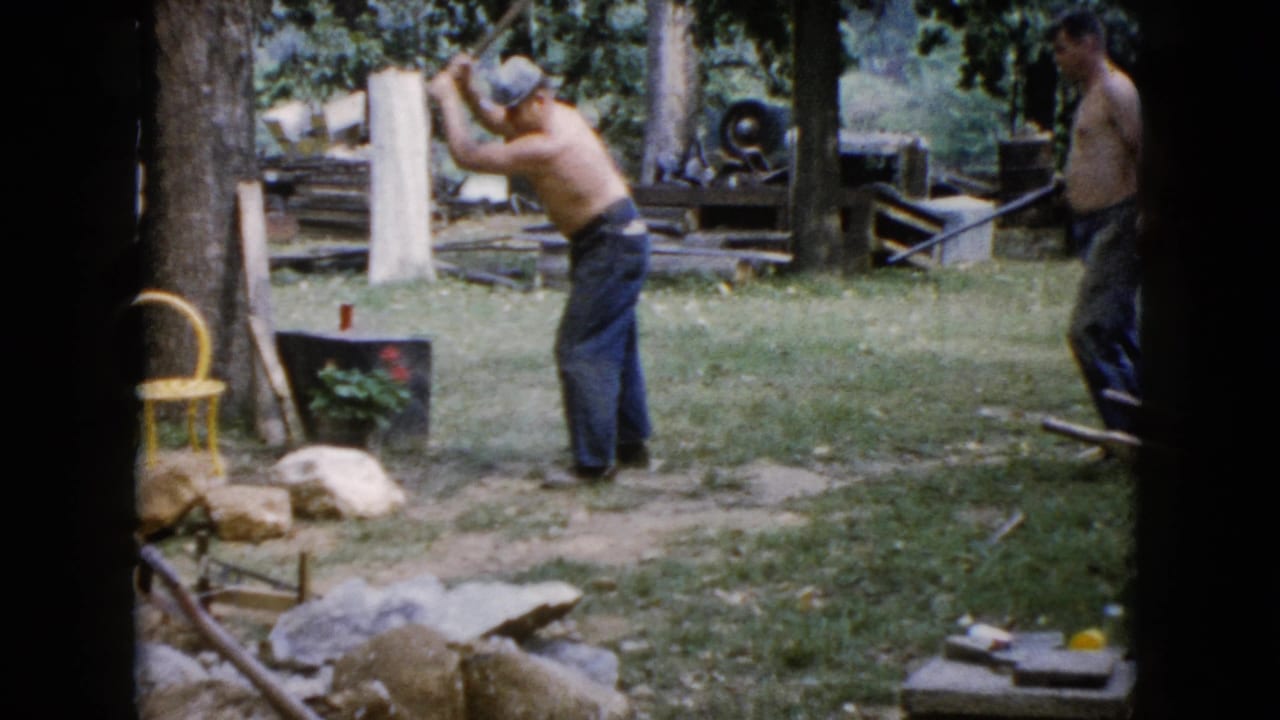 A shirtless man raises a sledgehammer in a grassy Wauconda yard while another man stands nearby with a tool.