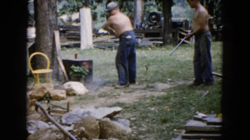 Two shirtless men work with hand tools in a grassy Wauconda yard, with stones, trees, and a yellow chair in view.