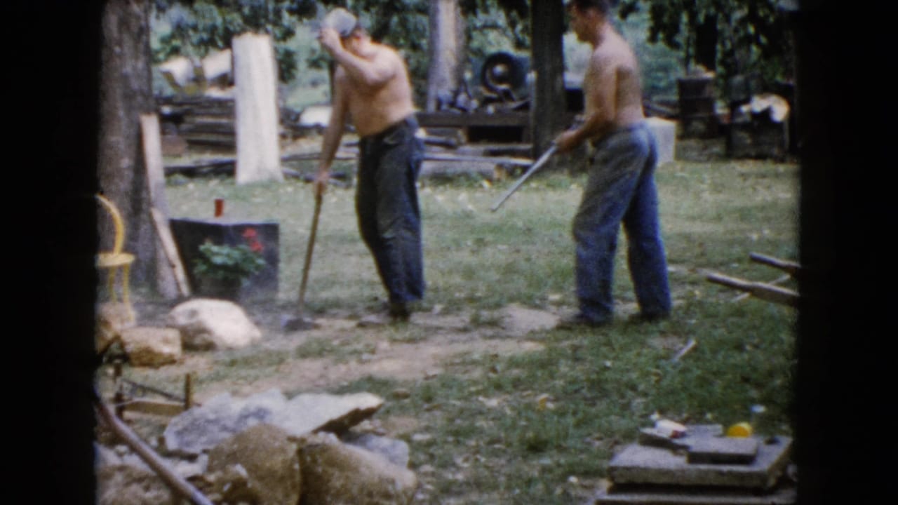 In 1960 Wauconda, Illinois home-movie footage, two shirtless men work outdoors with long-handled tools near rocks, trees, and a yard work area.