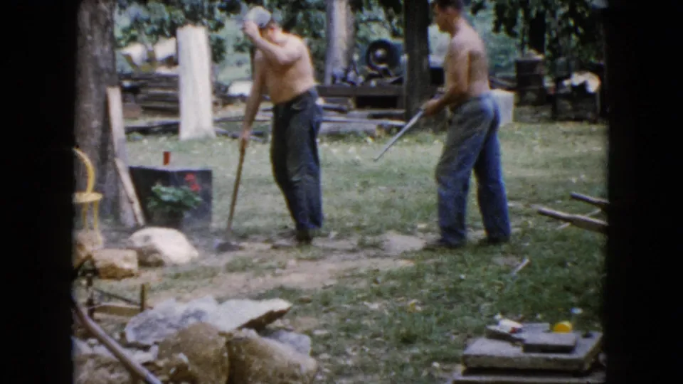 In 1960 Wauconda, Illinois home-movie footage, two shirtless men work outdoors with long-handled tools near rocks, trees, and a yard work area.