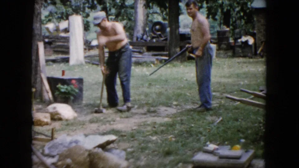 The 1960 home-movie frame shows one man bending with a hand tool while another stands nearby in a grassy Wauconda yard with rocks and trees.