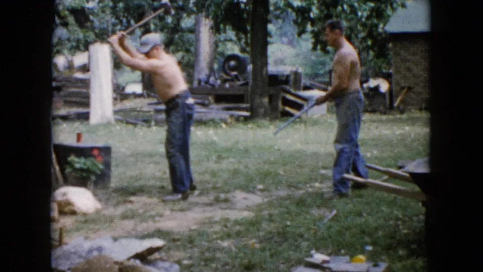 A shirtless man raises a long-handled tool in the grassy yard while another man stands nearby, with rocks, trees, and work materials visible.