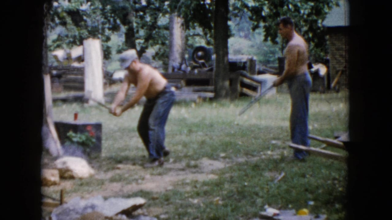 In 1960 Wauconda, Illinois home-movie footage, one shirtless man swings a sledgehammer at a rock while another watches with a tool in a tree-lined yard.