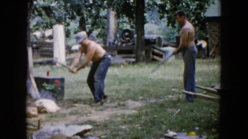 In 1960 Wauconda, Illinois home-movie footage, one shirtless man swings a sledgehammer at a rock while another watches with a tool in a tree-lined yard.