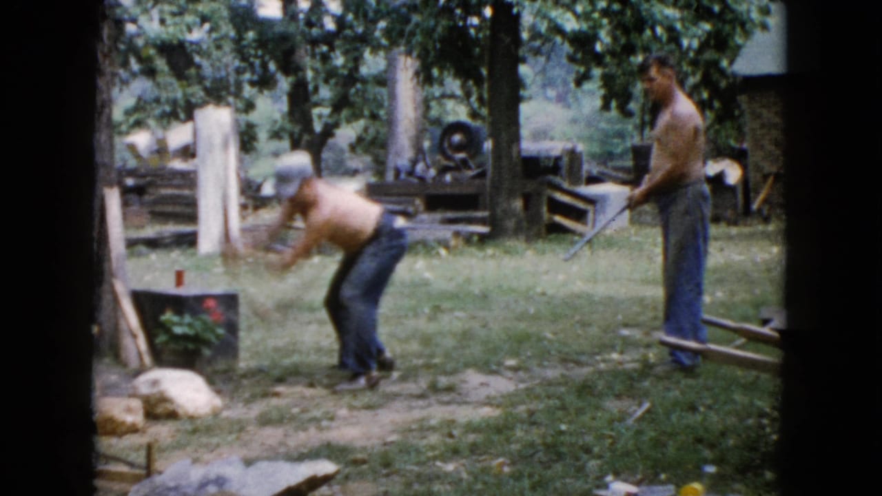 One man bends forward swinging a sledgehammer at a rock while another holds a long-handled tool in the yard.