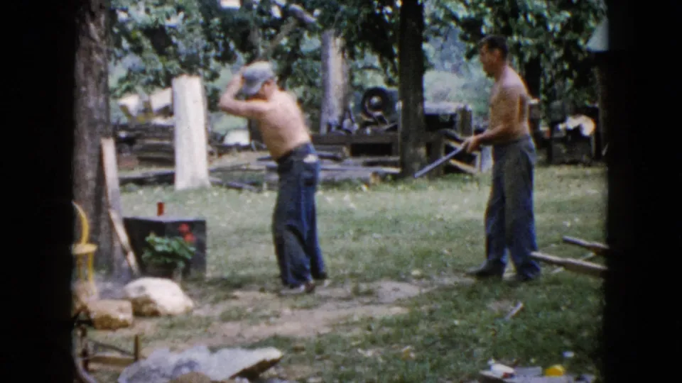 In the Wauconda home-movie frame, the hammering man lifts the sledgehammer for another strike as the second man watches from the grass.