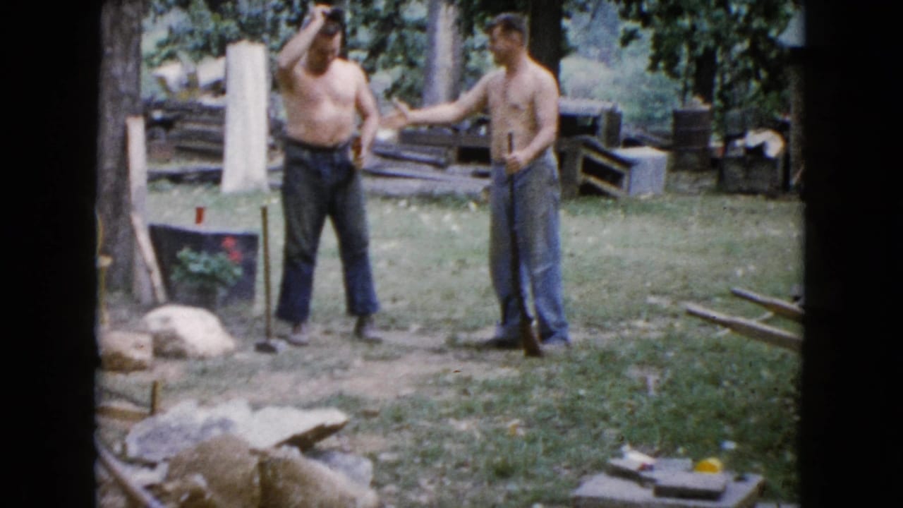 Two shirtless men stand in a grassy yard with a sledgehammer and scattered rocks nearby.