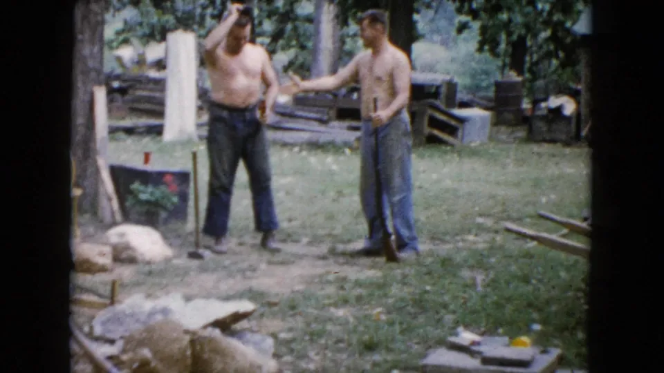 In 1960 Wauconda, Illinois, two shirtless men stand in a backyard work area near trees, lumber, rocks, and a sledgehammer.