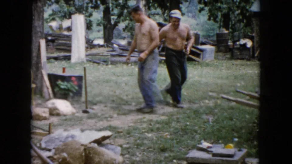 A shirtless man moves through the Wauconda backyard holding a tool as another man follows, with lumber and rocks visible around them.