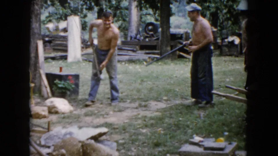 In the backyard scene, one shirtless man handles a sledgehammer while another man holds a long rifle-like object near trees and lumber.