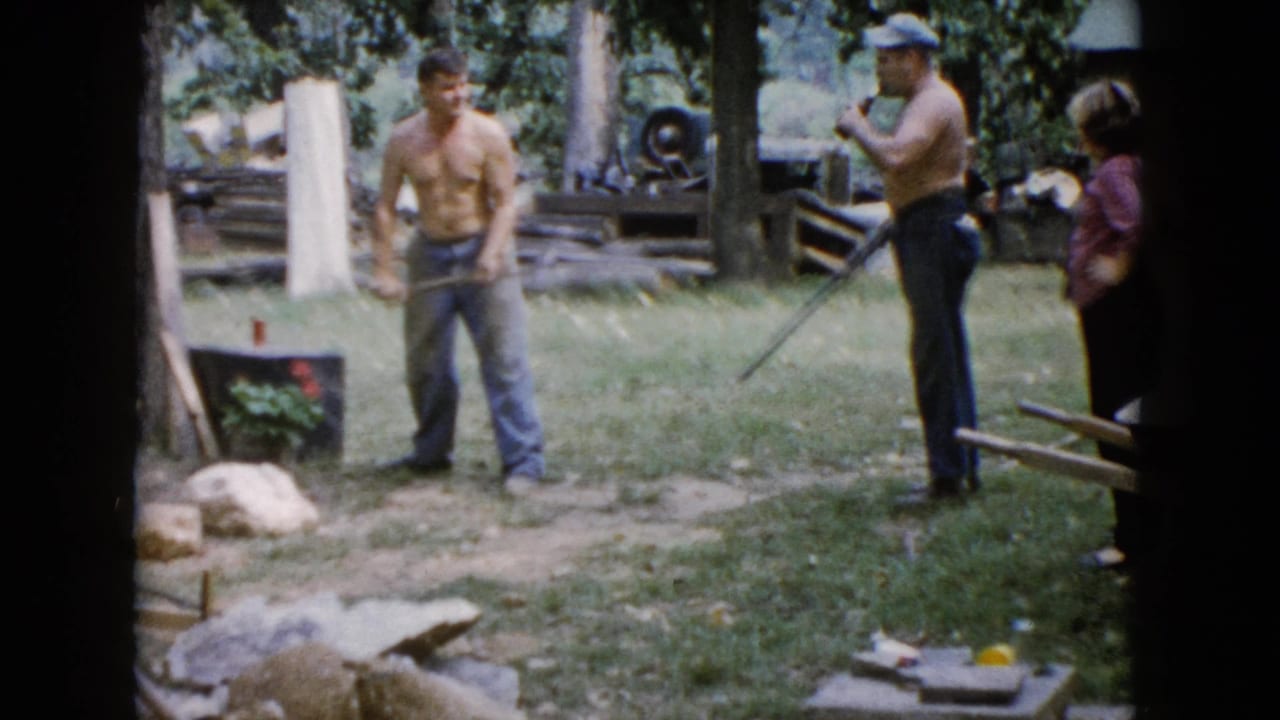 A shirtless man pauses with a sledgehammer in a Wauconda yard while another shirtless man in a cap and a woman stand beside him.