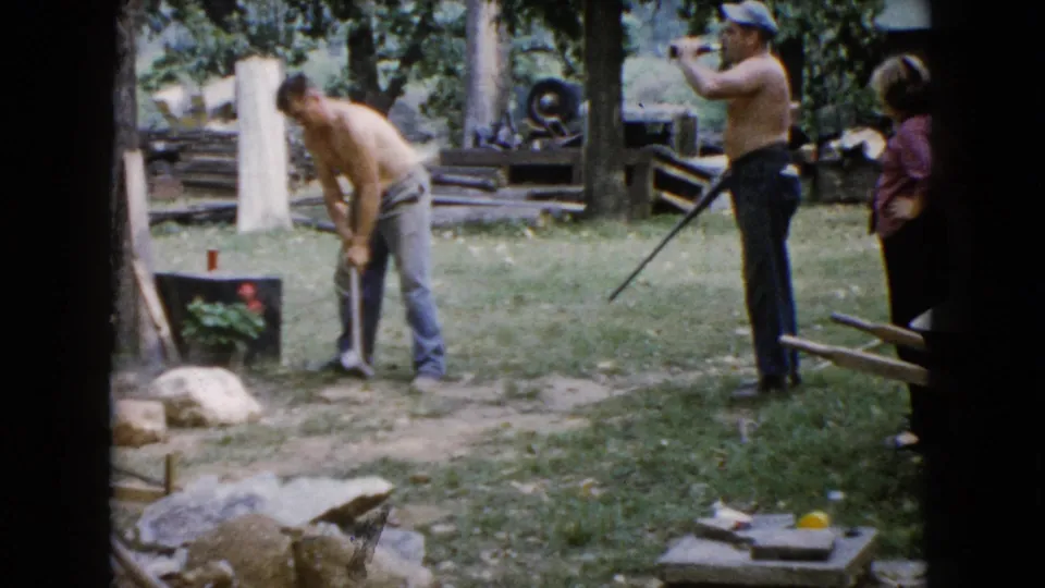 The shirtless man bends toward a rock with the sledgehammer in the 1960 Wauconda yard scene while two adults remain nearby.