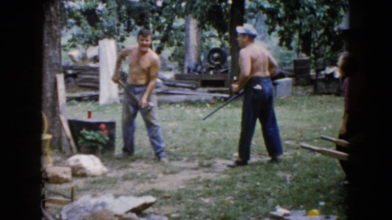 In a 1960 Wauconda, Illinois home-movie frame, two shirtless men stand in a shaded yard with an axe, rifle, logs, rocks, and a small planter nearby.