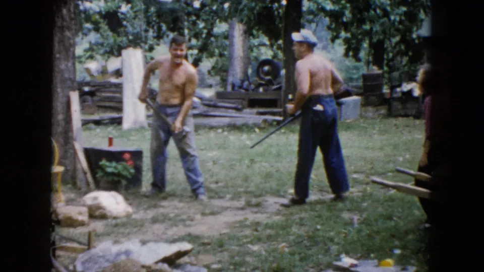 In a 1960 Wauconda, Illinois home-movie frame, two shirtless men stand in a shaded yard with an axe, rifle, logs, rocks, and a small planter nearby.