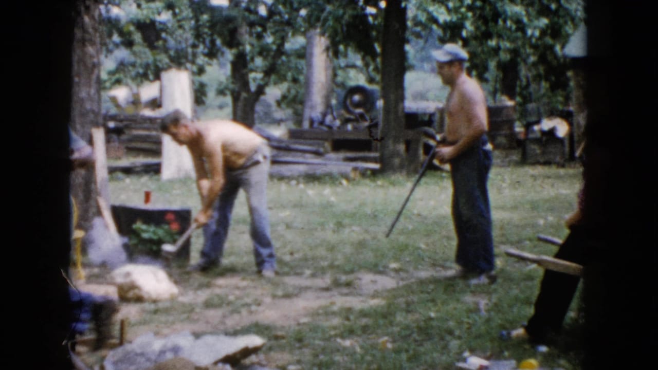 A 1960 Wauconda backyard frame shows one shirtless man bending with an axe as another shirtless man in a cap stands with a rifle near logs and trees.
