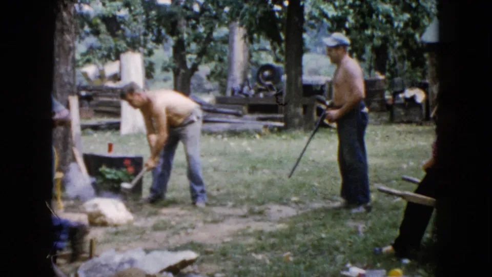A 1960 Wauconda backyard frame shows one shirtless man bending with an axe as another shirtless man in a cap stands with a rifle near logs and trees.
