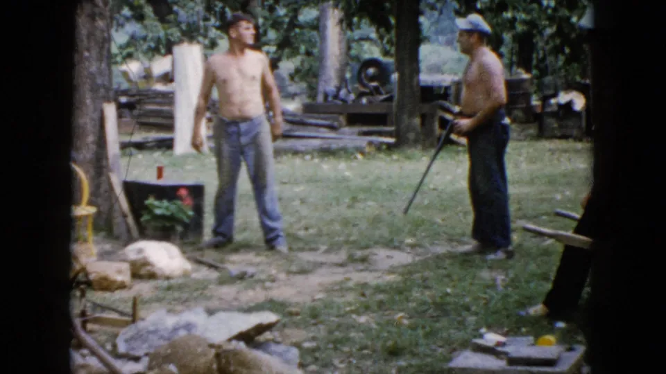 The 8mm frame shows two men in a Wauconda yard, one standing shirtless near rocks and a yellow chair while the other holds a rifle at his side.