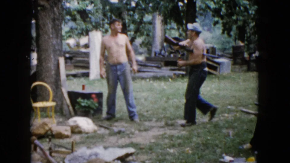 In the final 1960 Wauconda frame, a shirtless man stands in the grass while a capped man steps past holding a rifle, with logs, trees, and a yellow chair visible.