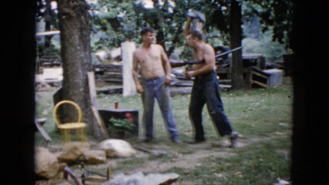 Two shirtless men stand in a grassy backyard as one holds a long gun near a yellow chair.