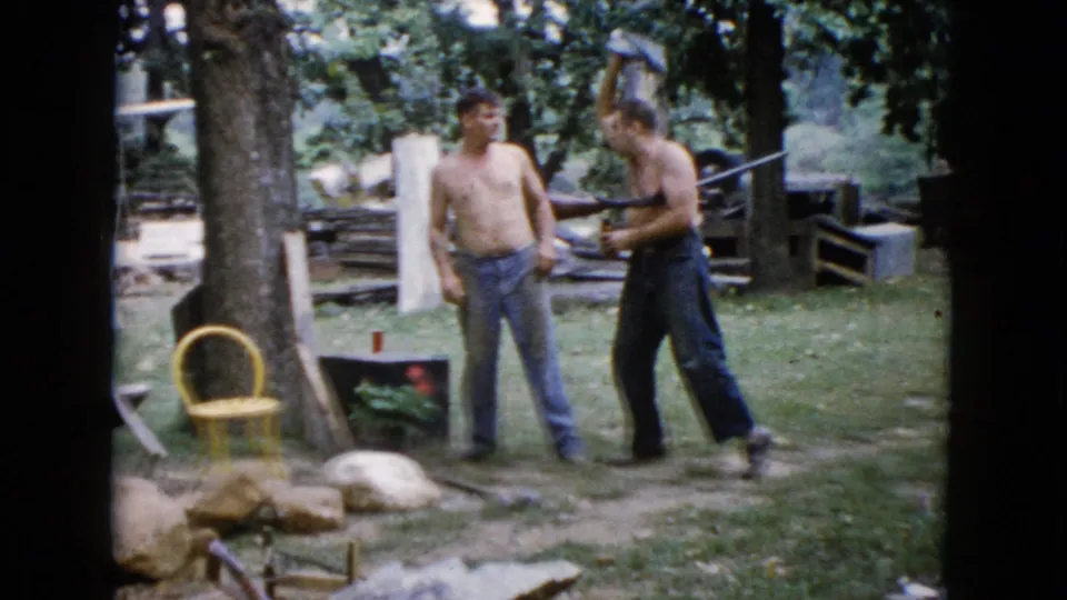 In Wauconda, Illinois around 1960, two shirtless men stand outdoors while one holds a long gun beside trees and yard objects.