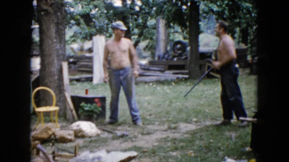 A 1960 Wauconda home-movie frame shows one man holding a long gun at waist level as another watches in the yard.