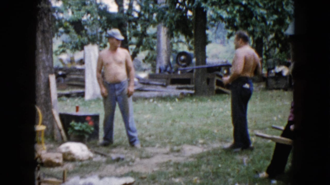 Two shirtless men stand in a grassy yard, one holding a long gun near trees and rocks.