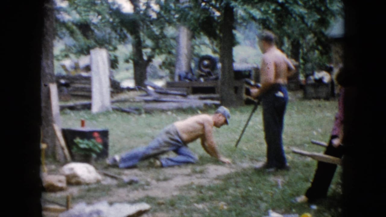 A 1960 Wauconda backyard scene shows one shirtless man dropping toward the grass while another stands nearby with a rifle-like long gun.