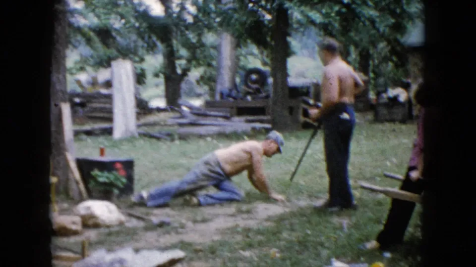 A 1960 Wauconda backyard scene shows one shirtless man dropping toward the grass while another stands nearby with a rifle-like long gun.