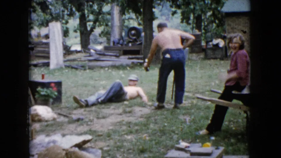In the 1960 Wauconda yard footage, a shirtless man lies on the grass while another man stands nearby and a woman in pink watches from a bench.