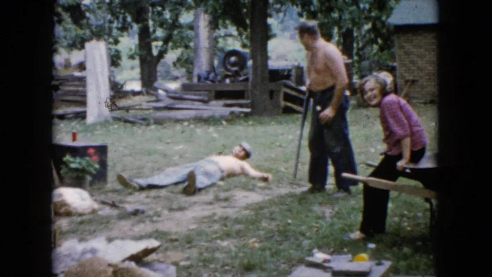 The 1960 Wauconda home movie shows a shirtless man lying on the lawn as another man stands near him and a woman in pink smiles beside a bench.