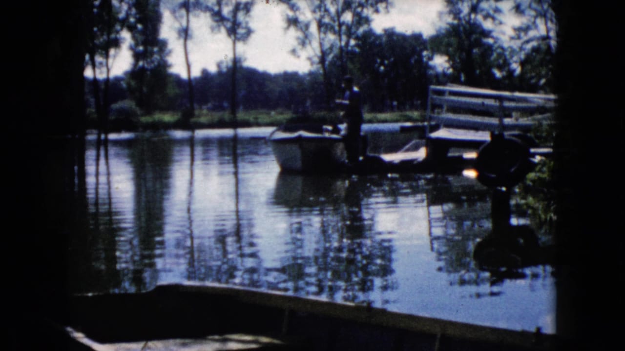 Man standing beside a small boat on calm lake water near a dock and trees.