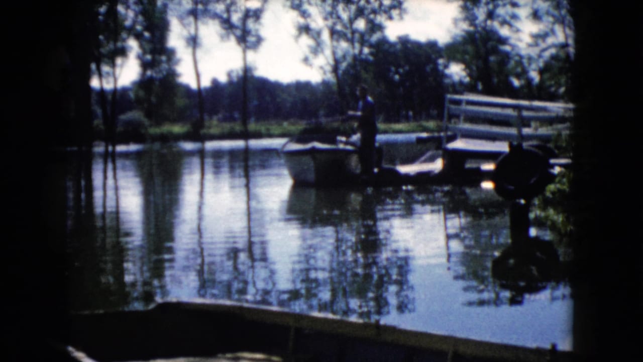 1960 Wauconda, Illinois home-movie frame of a man by a small boat near a dock on a calm, tree-lined lake.