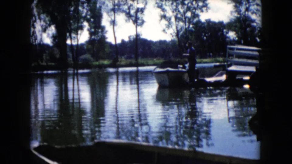 1960 Wauconda, Illinois 8mm view of a man beside a small boat, framed by lake reflections and a dock rail.