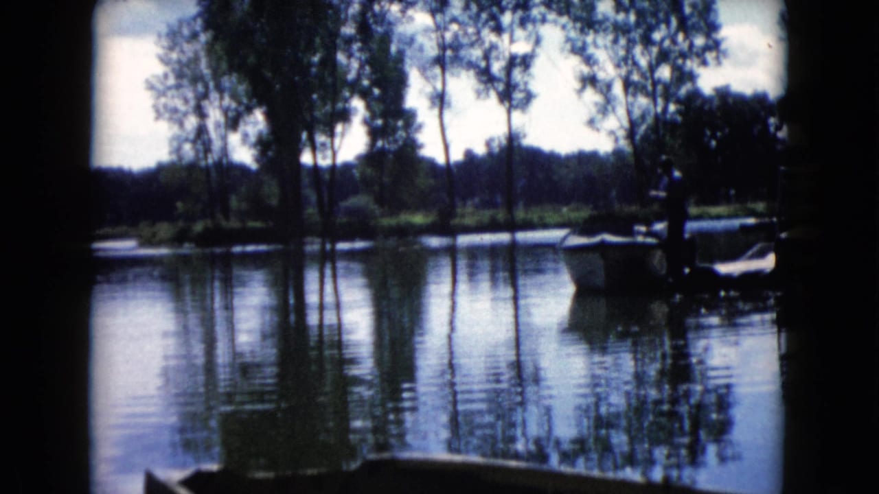 Man standing near a small boat on a calm lake with trees reflected in the water.