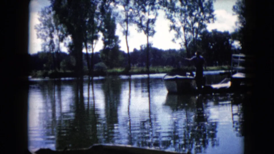 1960 home-movie view of a man near a small boat on a wooded Wauconda lake, with soft focus and film grain.