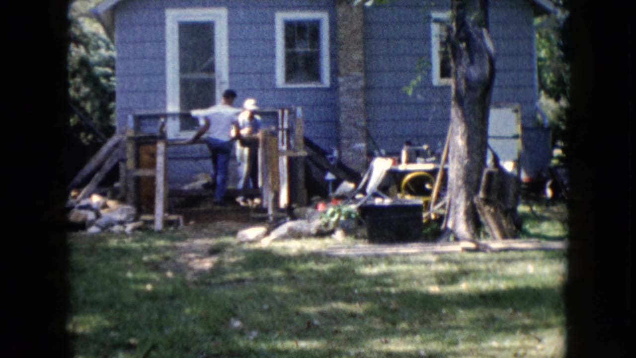 Two men work on a wooden structure behind a blue house in a backyard.