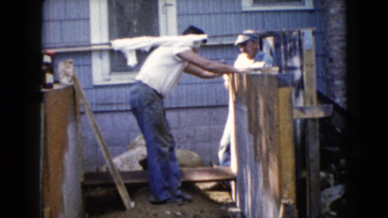 A man leans over a wooden frame while another man stands behind the boards.