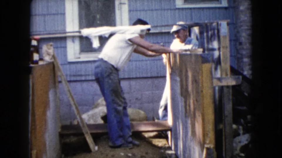 Two men work closely on a wooden porch frame beside a blue house, with one leaning forward and the other partly hidden by boards.
