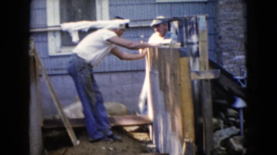 A close home-movie view shows a man in jeans leaning over porch framing as another man stands behind upright boards near the house.