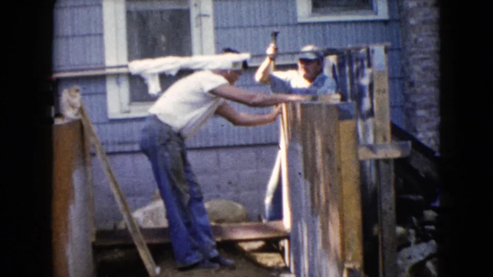 A man lifts a hammer over the wood framing while another leans in to hold or align the porch structure beside the blue house.