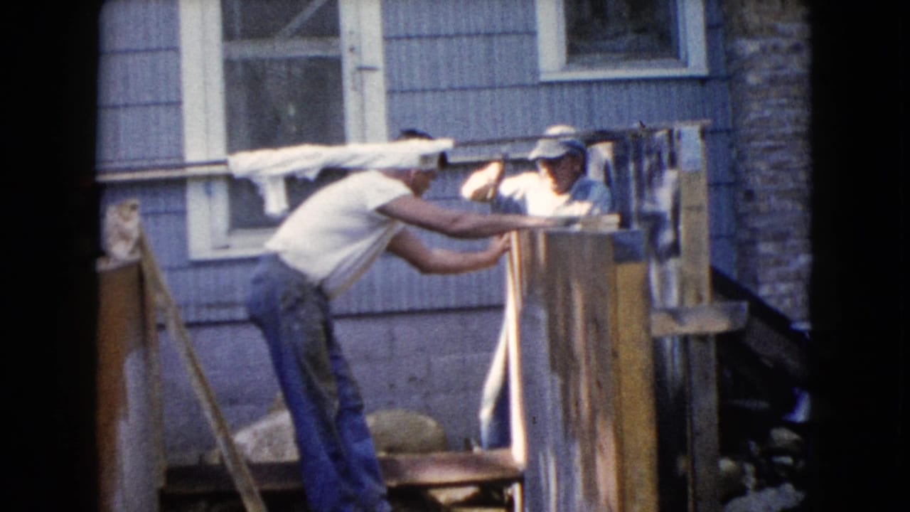 Two men work on a wooden structure outside a shingled house.